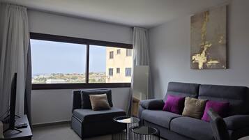 A modern living room with a grey sofa, a black armchair, and a round coffee table. A large window offers a view of buildings and greenery.