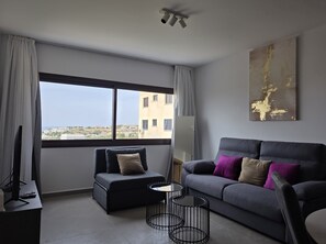 A modern living room with a grey sofa, a black armchair, and a round coffee table. A large window offers a view of buildings and greenery. - Kentrikon 1924 Boutique Hotel And Suites (Paphos)