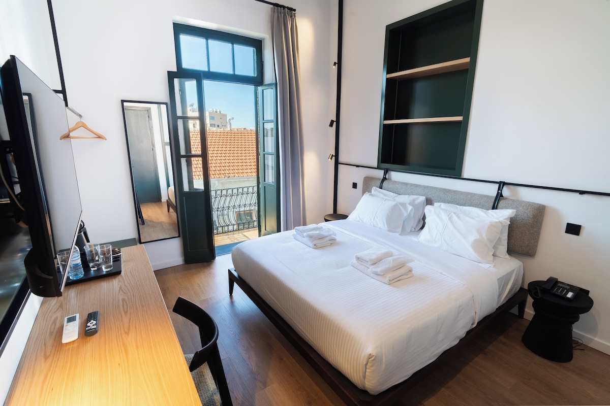 A hotel room with a bed, bedside table, desk, chair, and a view of the outside.