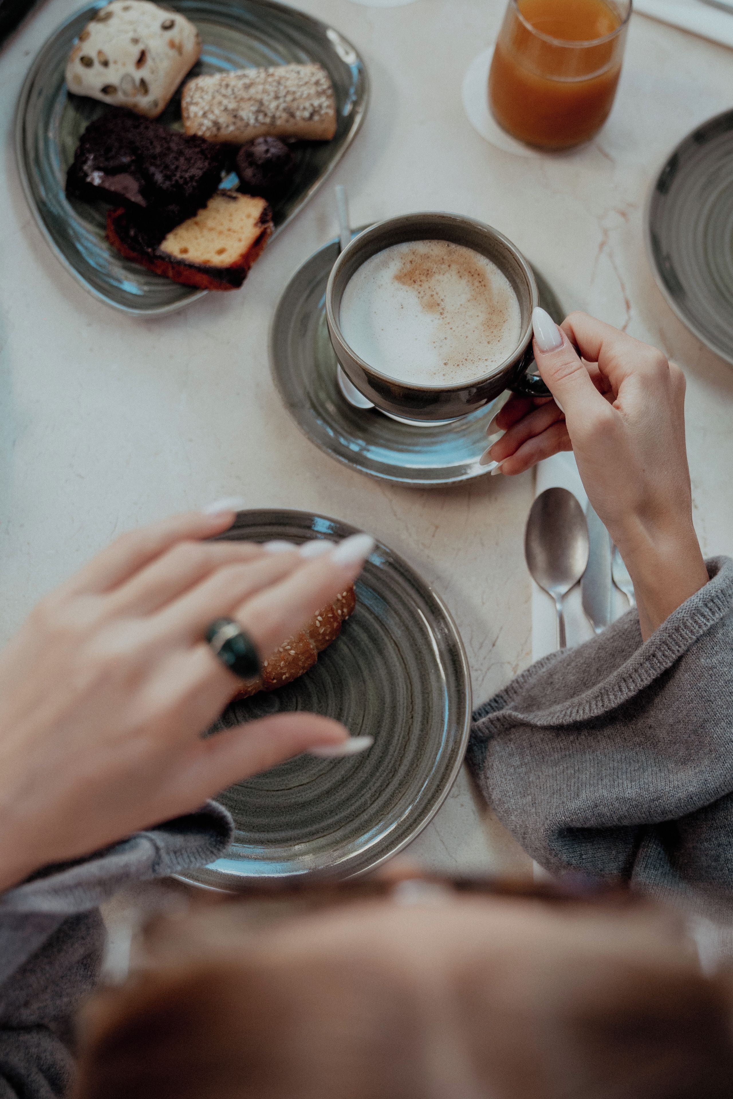 Une personne tenant une tasse de café avec une soucoupe, une assiette de biscuits et un verre de jus d’orange sur une table.