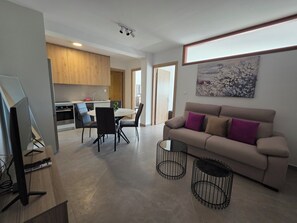A modern living room with a beige sofa, a dining area with a table and chairs, a kitchen with wooden cabinets, and a painting on the wall. - Kentrikon 1924 Boutique Hotel And Suites (Paphos)