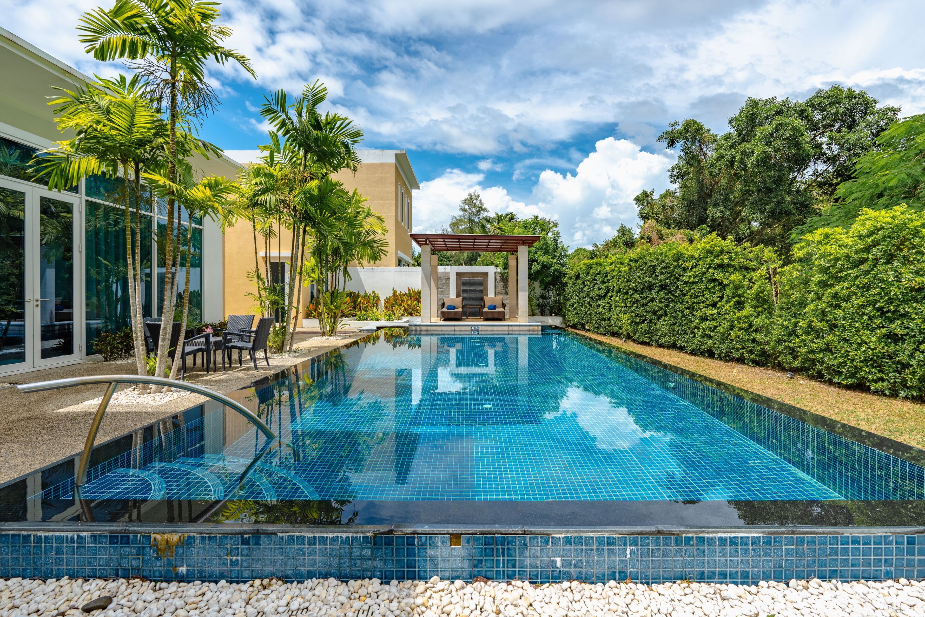 Piscina com pergolado, palmeiras e um edifício moderno com portas de vidro.