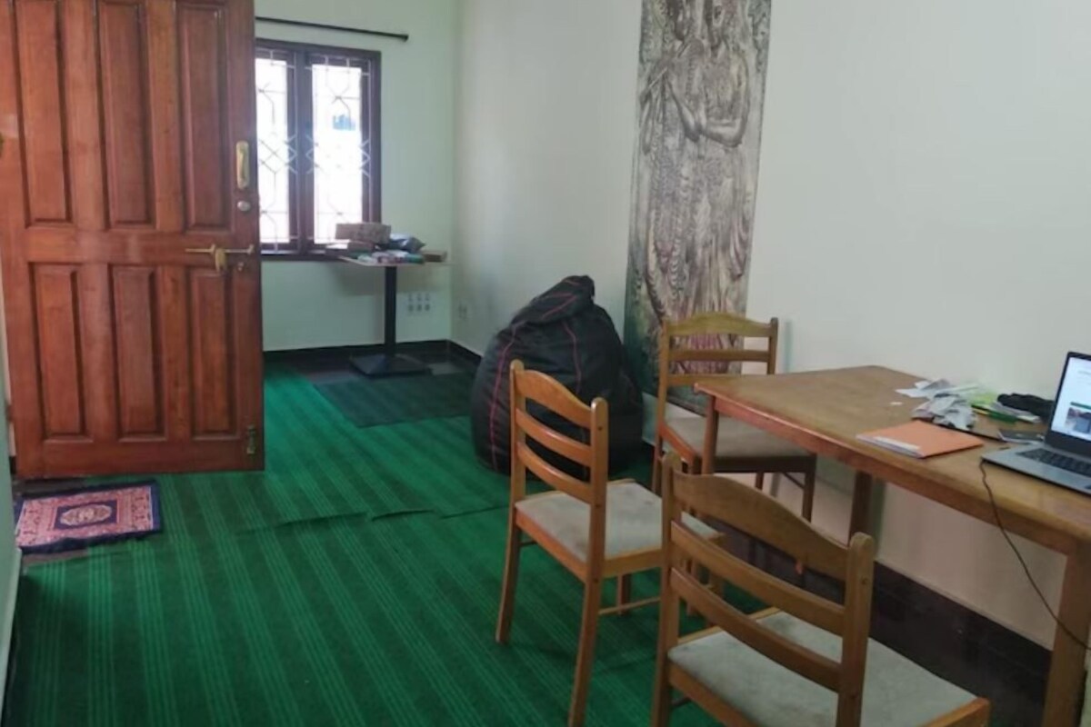 A room with a wooden door, a green carpet, a table with a laptop, and a chair.