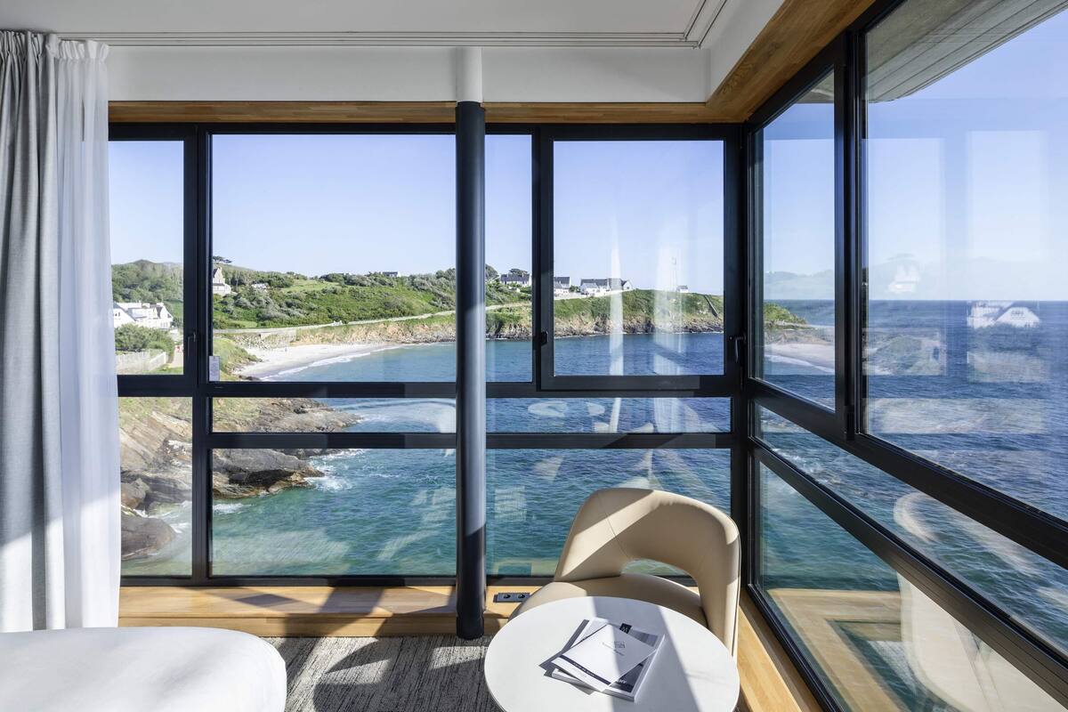 A room with large windows offering a coastal view, a small round table with a chair, and a bed.