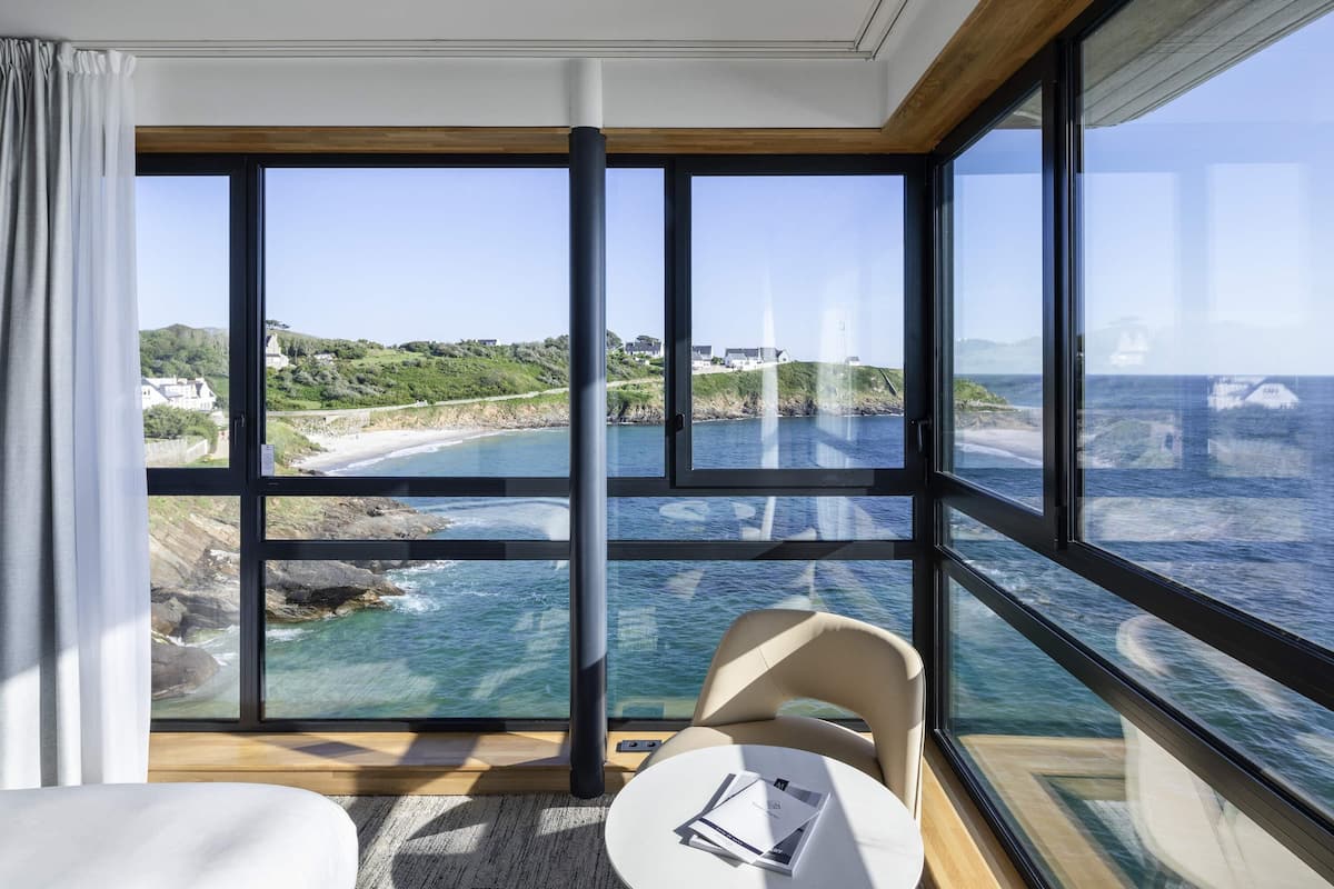 A room with large windows offering a coastal view, a small round table with a chair, and a bed.