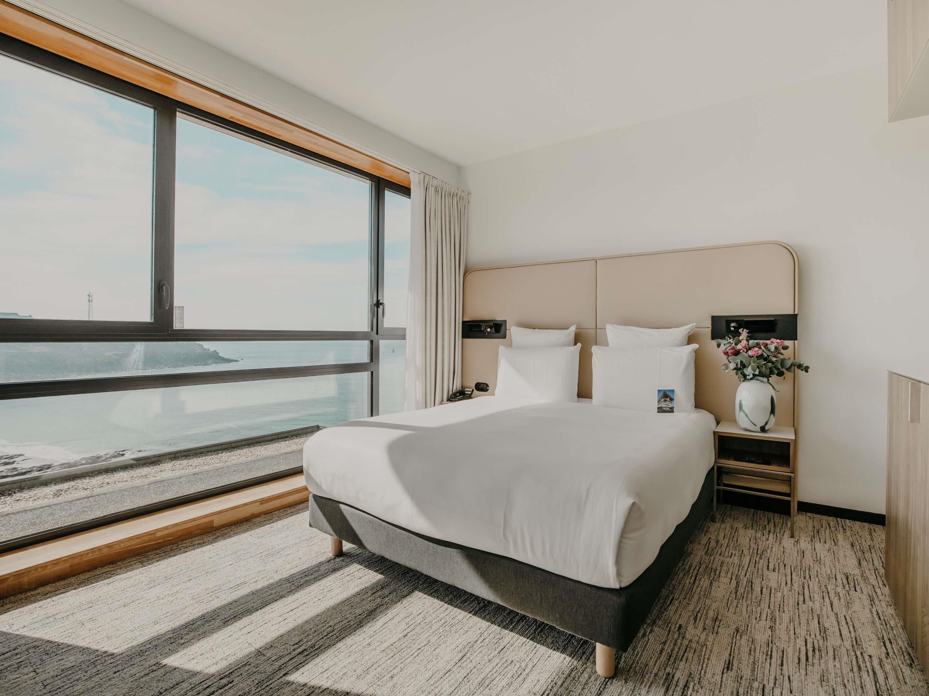 A hotel room with a large bed, a nightstand, a vase with flowers, and a view of the ocean.