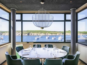 Un coin repas avec une table dressée pour six personnes, de grandes fenêtres offrant une vue sur les bateaux et les falaises, et un luminaire suspendu.