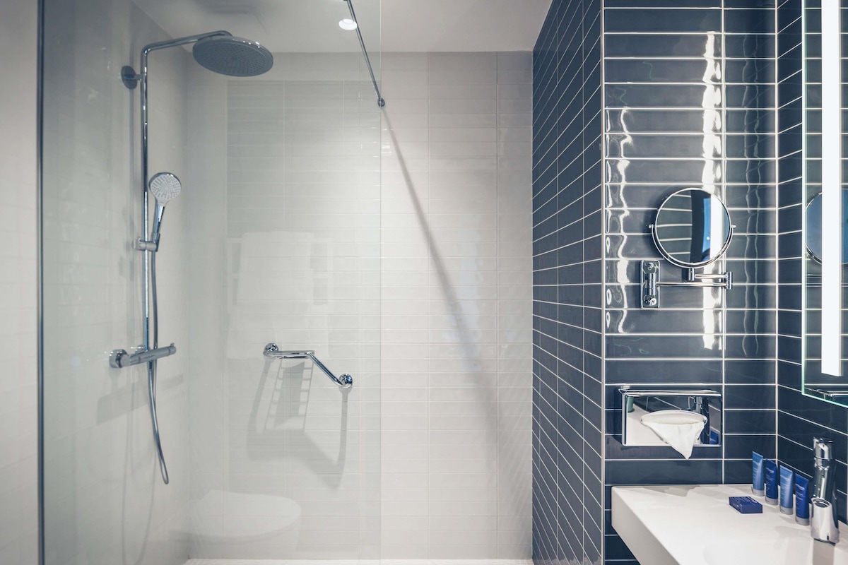 Un baño moderno con cabina de ducha de vidrio, un lavabo blanco y una pared revestida de baldosas oscuras.