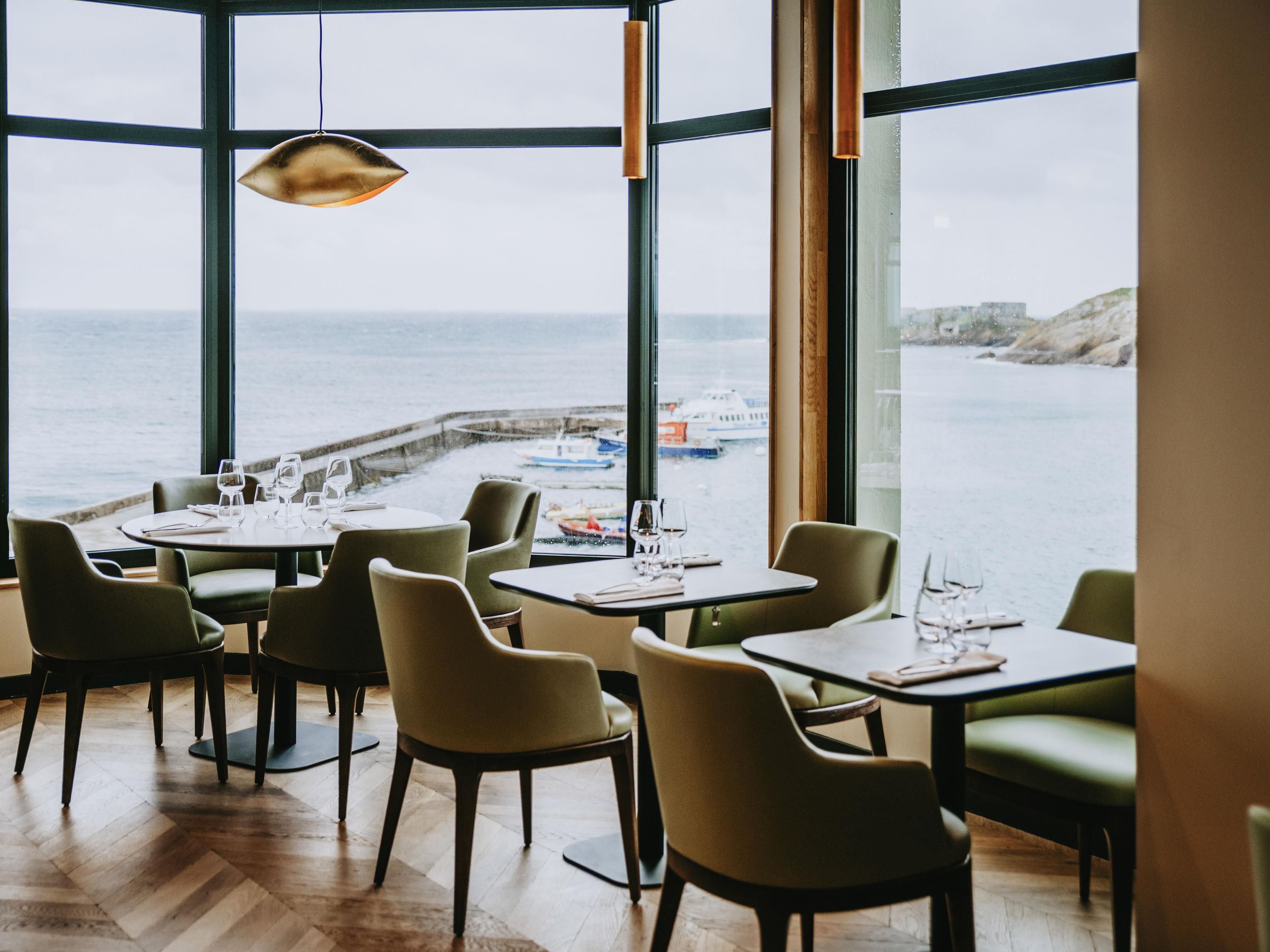 A restaurant with tables and chairs, large windows with a view of the sea and boats.