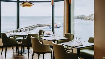 A restaurant with tables and chairs, large windows with a view of the sea and boats.