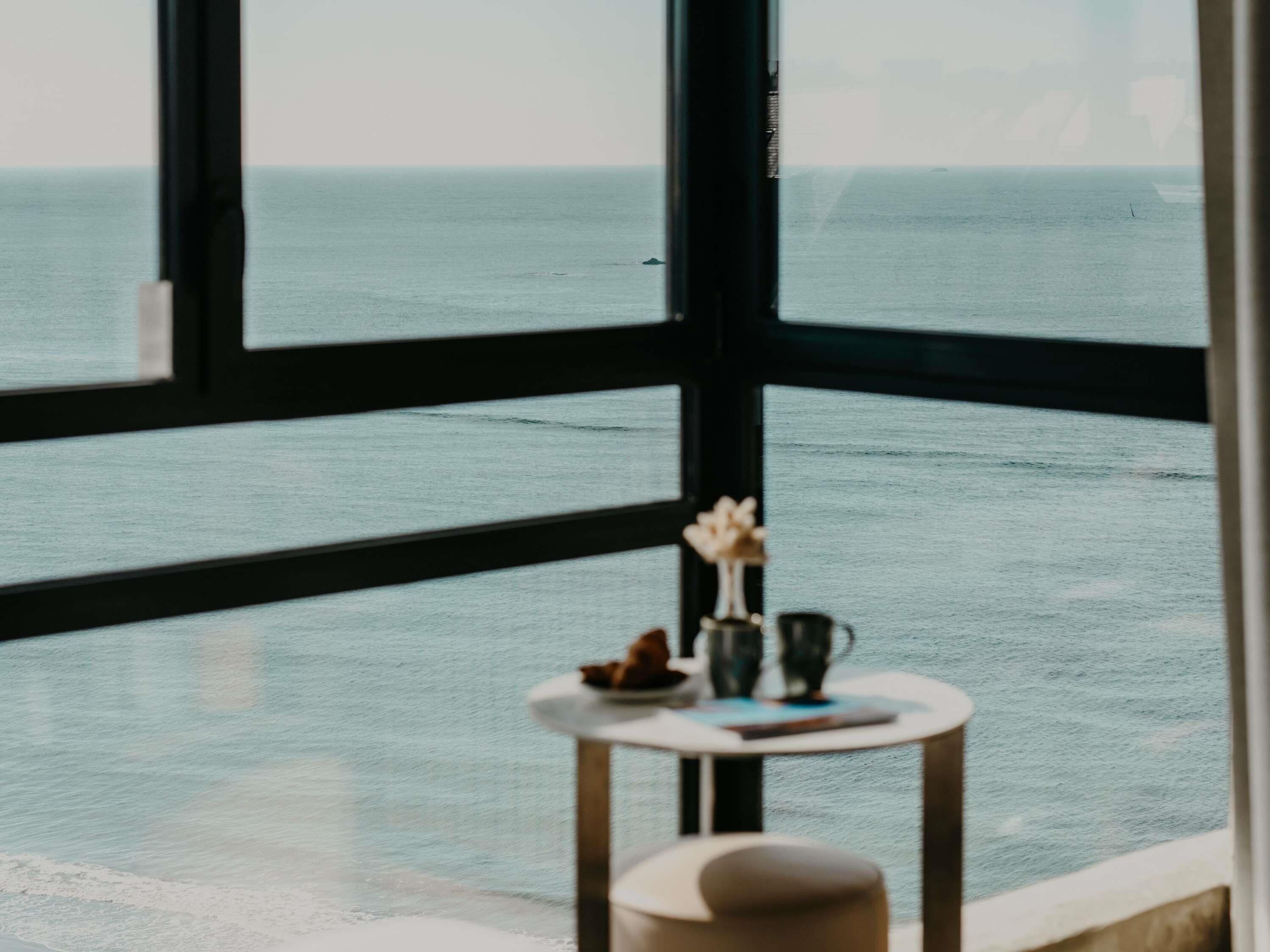 Een kamer met een groot raam dat uitkijkt op zee, een klein rond tafeltje met een vaas en een boek, en een stoel.
