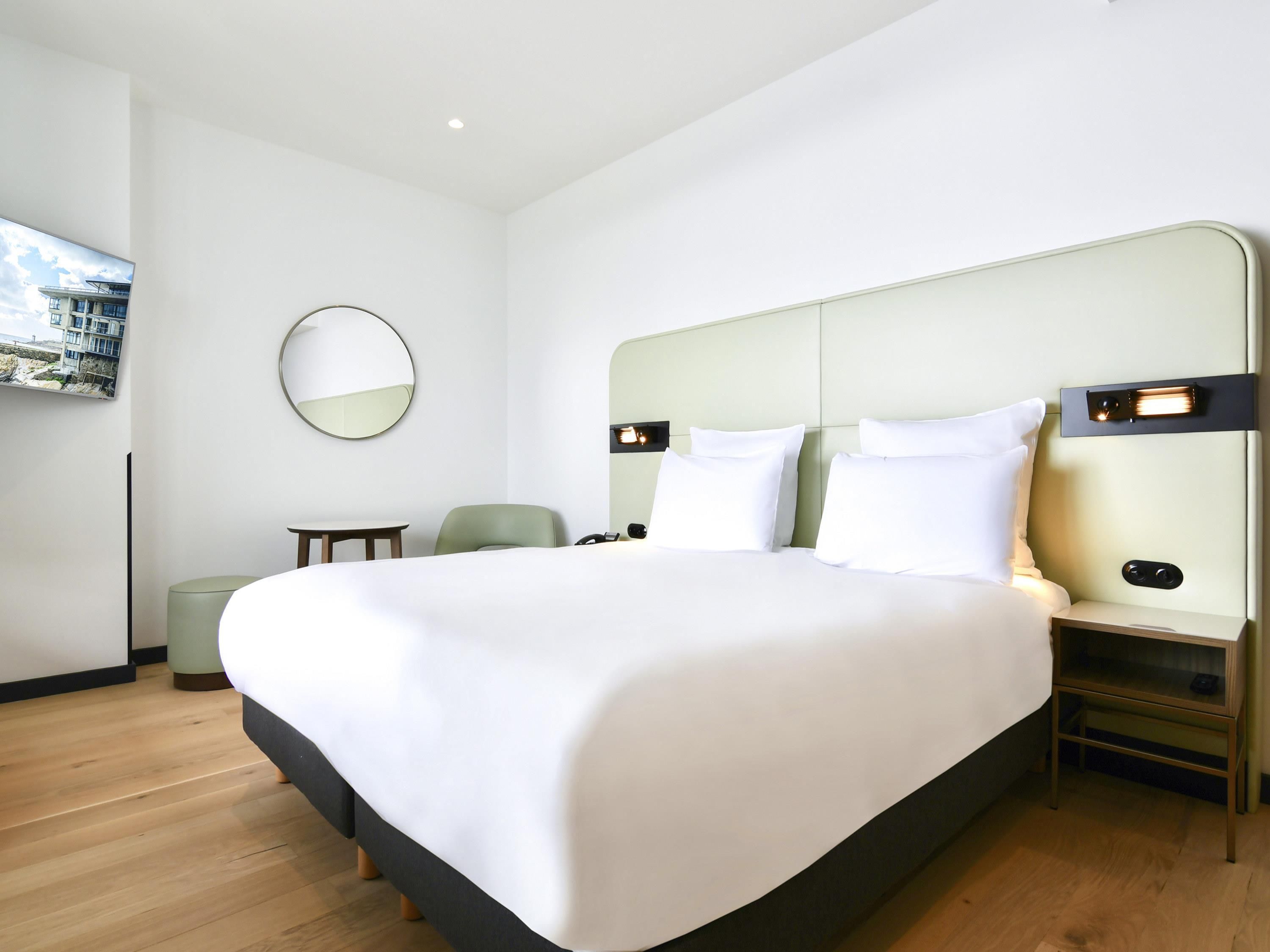 A modern hotel room with a large bed, a bedside table, a chair, a round mirror, and a wall-mounted TV.