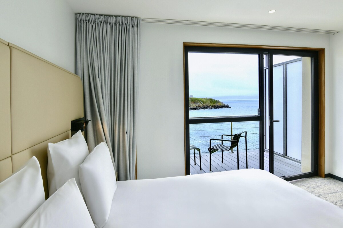 A hotel room with a bed, a view of the sea through a large window, and a balcony with a chair.