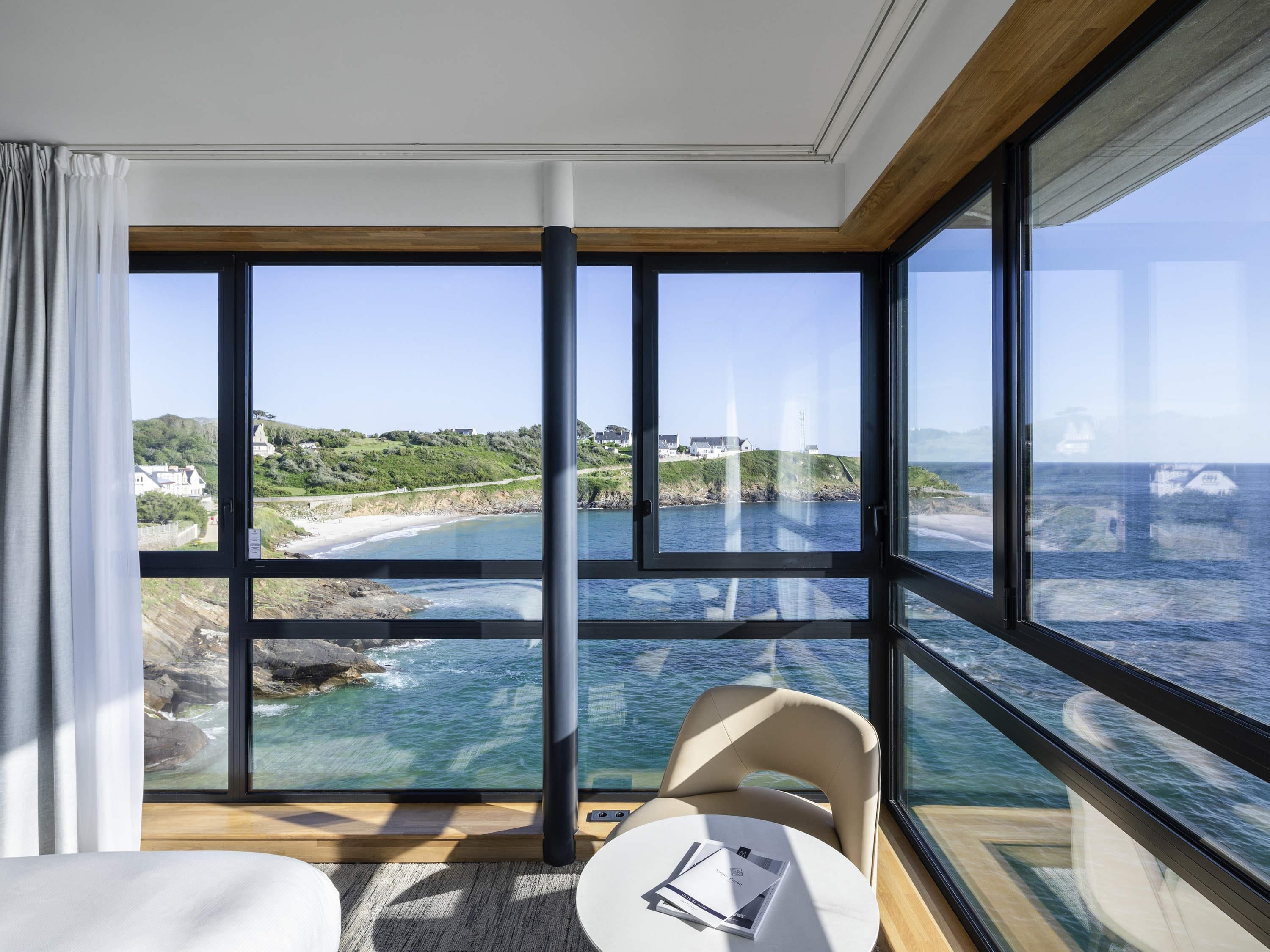 Ein Zimmer mit großen Fenstern und Blick auf die Küste, ein kleiner runder Tisch mit einem Stuhl und ein Bett.