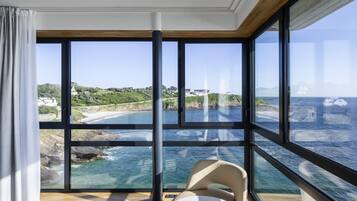 Ein Zimmer mit großen Fenstern und Blick auf die Küste, ein kleiner runder Tisch mit einem Stuhl und ein Bett.