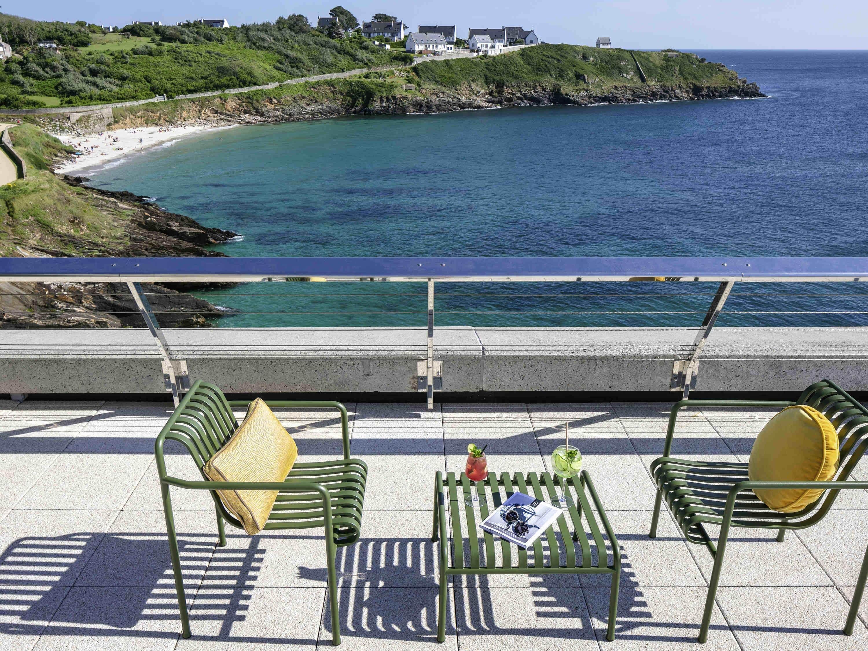 Une terrasse en bord de mer avec des chaises en métal vert, une table avec des boissons et une vue sur l’océan et les maisons situées sur une colline.