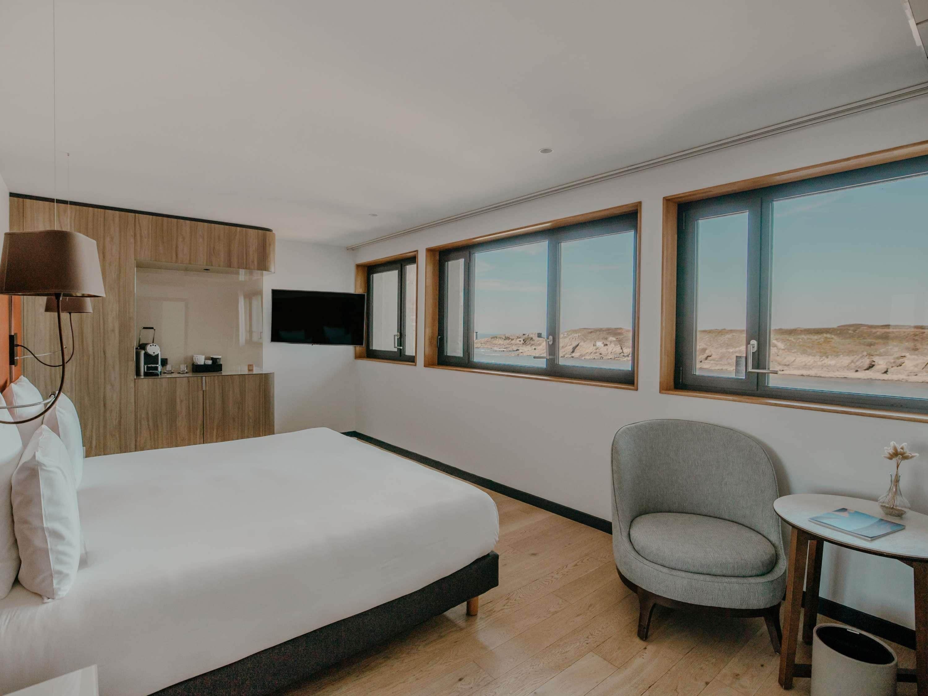 A bedroom with a bed, a chair, a small table, a TV, and large windows with a beach view.