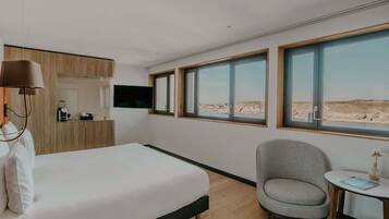 A bedroom with a bed, a chair, a small table, a TV, and large windows with a beach view.
