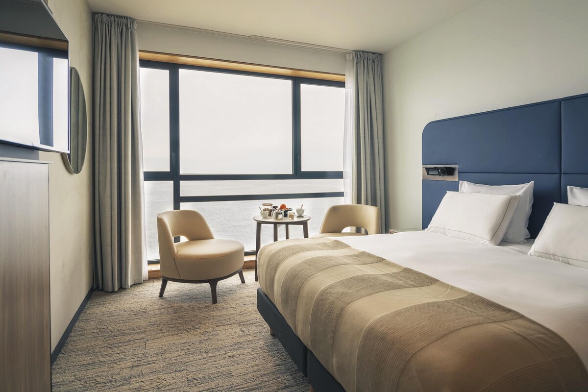 Una habitación de hotel con una cama grande, dos sillas, una mesita y un ventanal con cortinas.