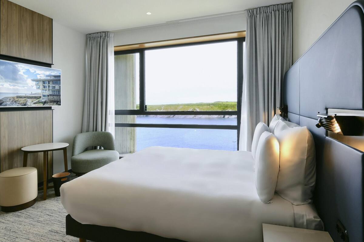 A hotel room with a large bed, a small round table, a chair, a TV mounted on the wall, and a view of the sea.