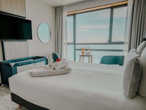 A hotel room with a large bed, a TV, a round mirror, a small table with a laptop, and a view of the sea.