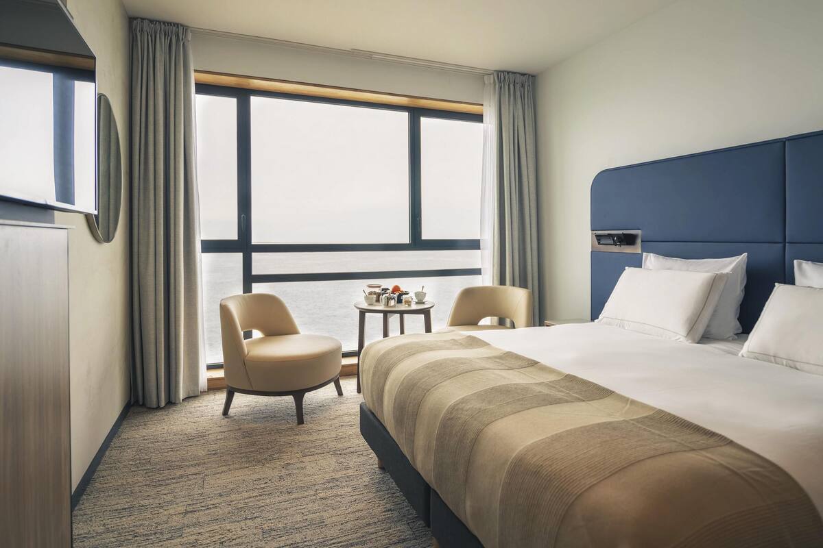 A hotel room with a large bed, two chairs, a small table, and a window with curtains.