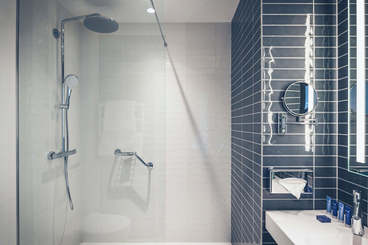 Un baño moderno con cabina de ducha de vidrio, un lavabo blanco y una pared revestida de baldosas oscuras.