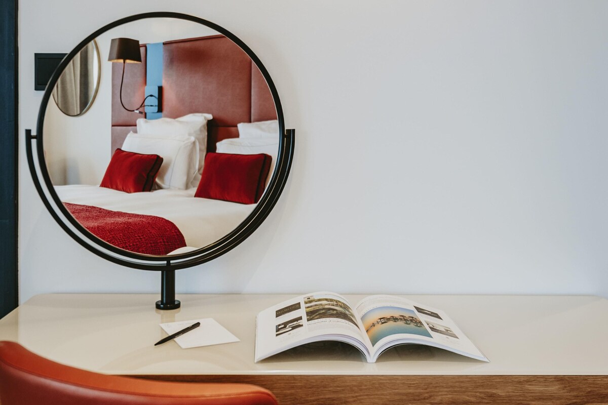 A hotel room with a bed, a round mirror, a wooden desk, and an open magazine.