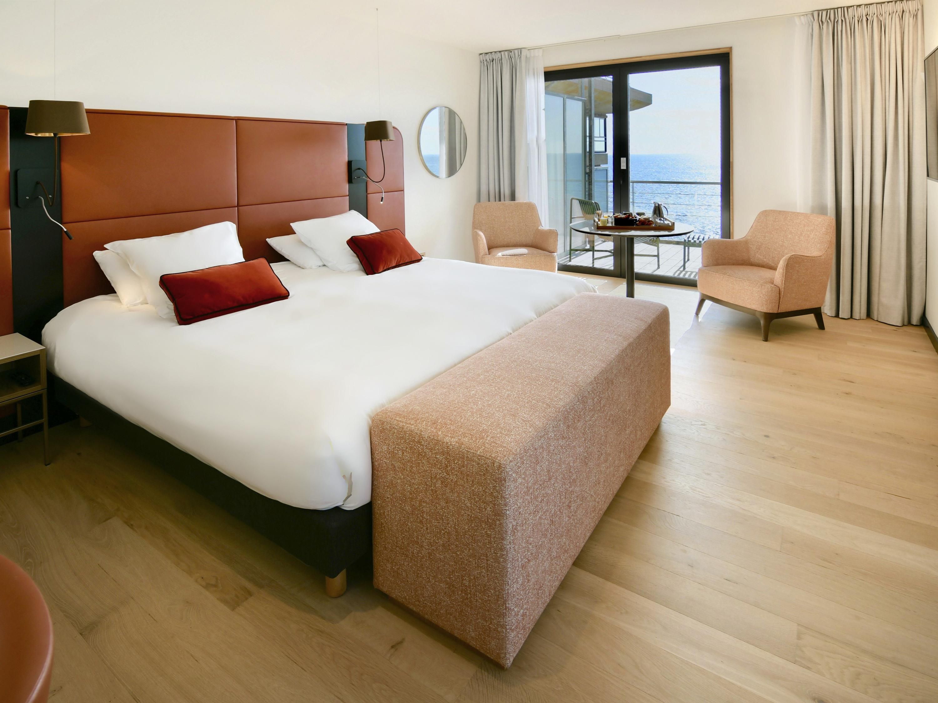 Una habitación de hotel moderna con una cama grande, una mesita de noche, una silla, una pequeña mesa con una bandeja y vistas al mar a través de un ventanal amplio.