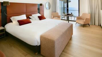 Una habitación de hotel moderna con una cama grande, una mesita de noche, una silla, una pequeña mesa con una bandeja y vistas al mar a través de un ventanal amplio.