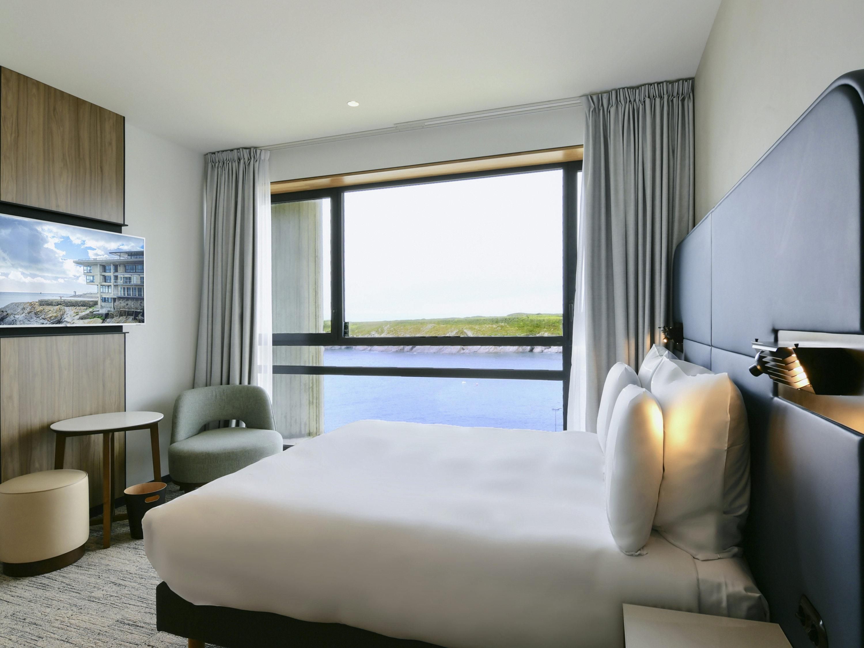 Ein Hotelzimmer mit einem großen Bett, einem kleinen runden Tisch, einem Sessel, einem an der Wand befestigten Fernseher und Meerblick.