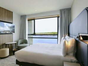 Habitación de hotel con una cama grande, una mesita redonda pequeña, una silla, un televisor montado en la pared y vista al mar.