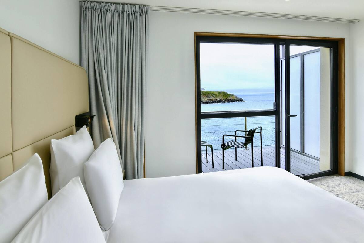 A hotel room with a bed, a view of the sea through a large window, and a balcony with a chair.