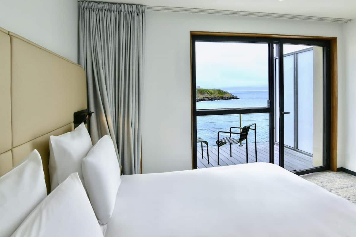 A hotel room with a bed, a view of the sea through a large window, and a balcony with a chair.