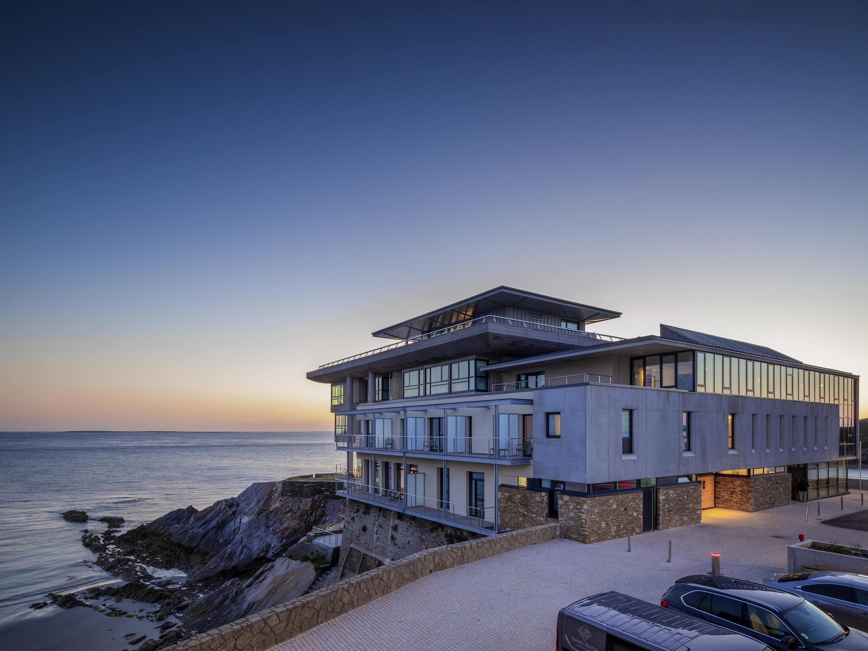 A modern building with multiple floors, large windows, and a balcony overlooking the sea.