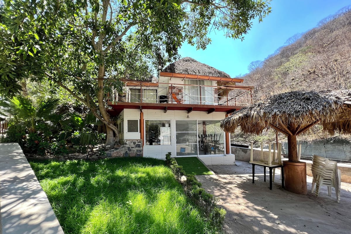 Una casa de dos pisos con un balcón, rodeada de vegetación exuberante y un tejado de paja.