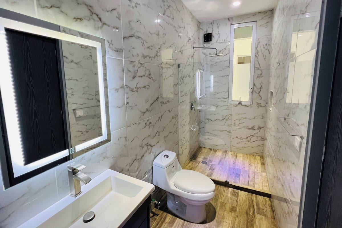 Un baño moderno con paredes de mármol, cabina de ducha de vidrio, inodoro blanco y un espejo grande.