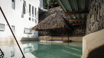 Un área de piscina con una estructura de techo de paja, paredes de piedra y un edificio blanco con ventanas.
