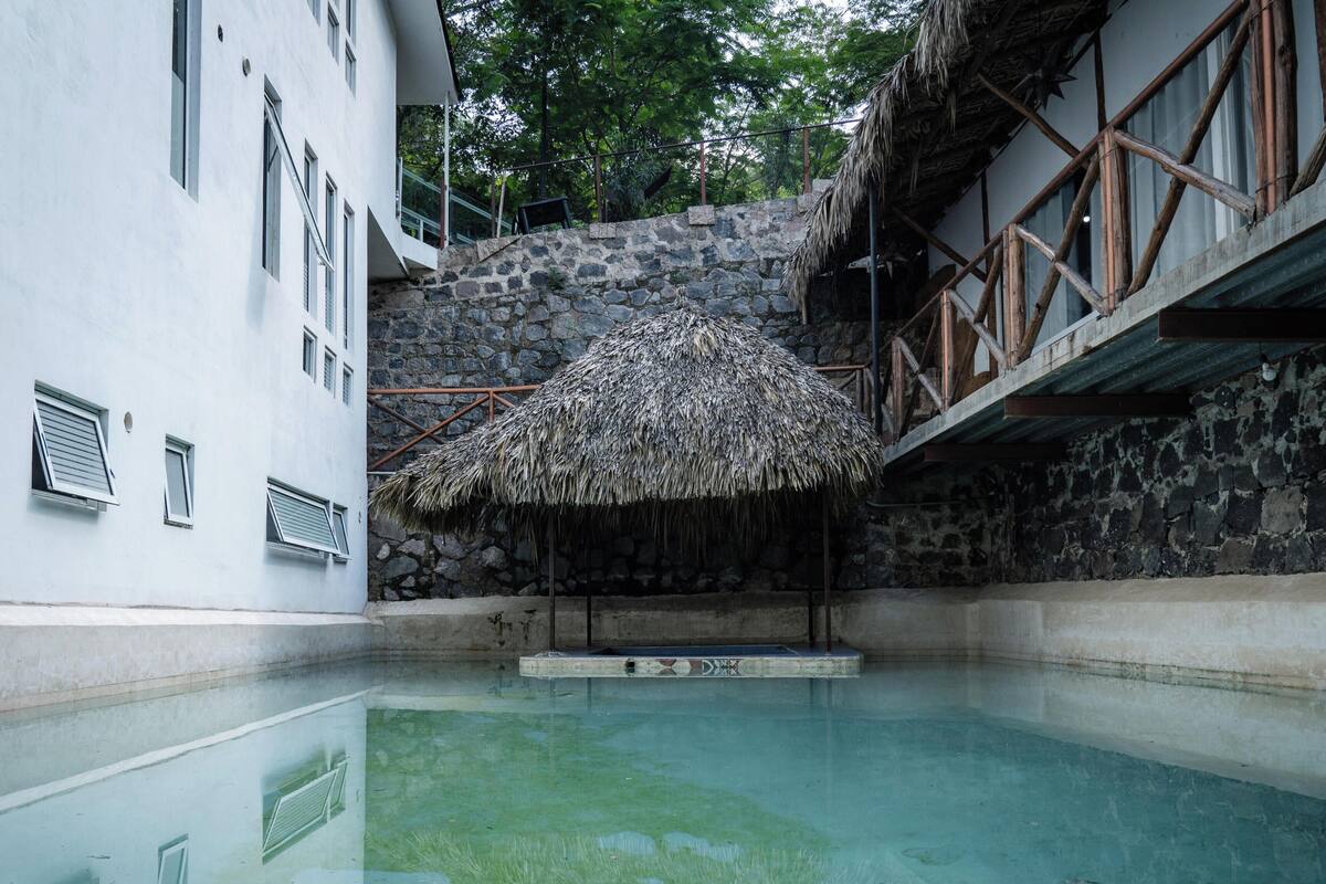 Una piscina con una estructura de techo de paja, paredes de piedra y un edificio blanco con ventanas.