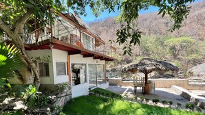 A house with a balcony, a thatched roof structure, and a garden with grass and trees.