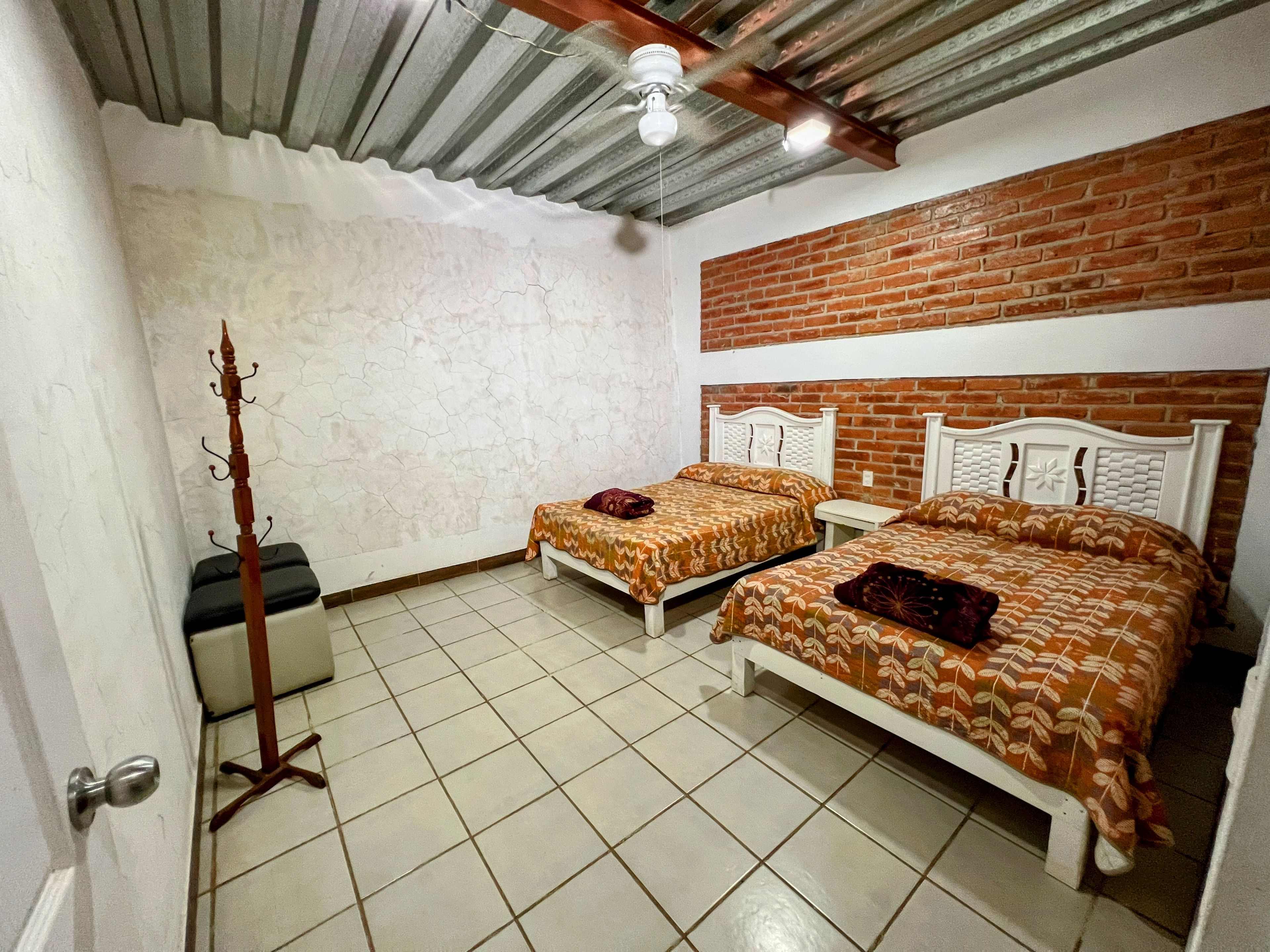 A room with two beds, a wooden coat rack, a ceiling fan, and a brick wall.