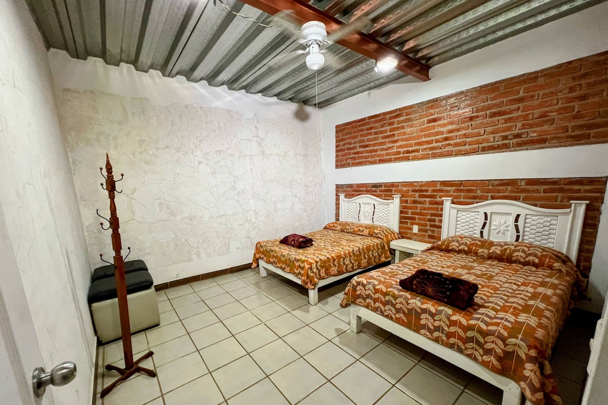 Ein Zimmer mit zwei Betten, einem Holz-Kleiderständer, einem Deckenventilator und einer Ziegelwand.