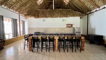 A spacious kitchen with a thatched roof, a central island with bar stools, and a tiled floor.