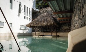 Un área de piscina con una estructura de techo de paja, paredes de piedra y un edificio blanco con ventanas.
