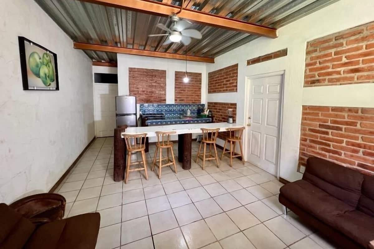 Una cocina con piso de baldosas, un refrigerador blanco, una mesa de madera con cuatro sillas y un ventilador de techo.