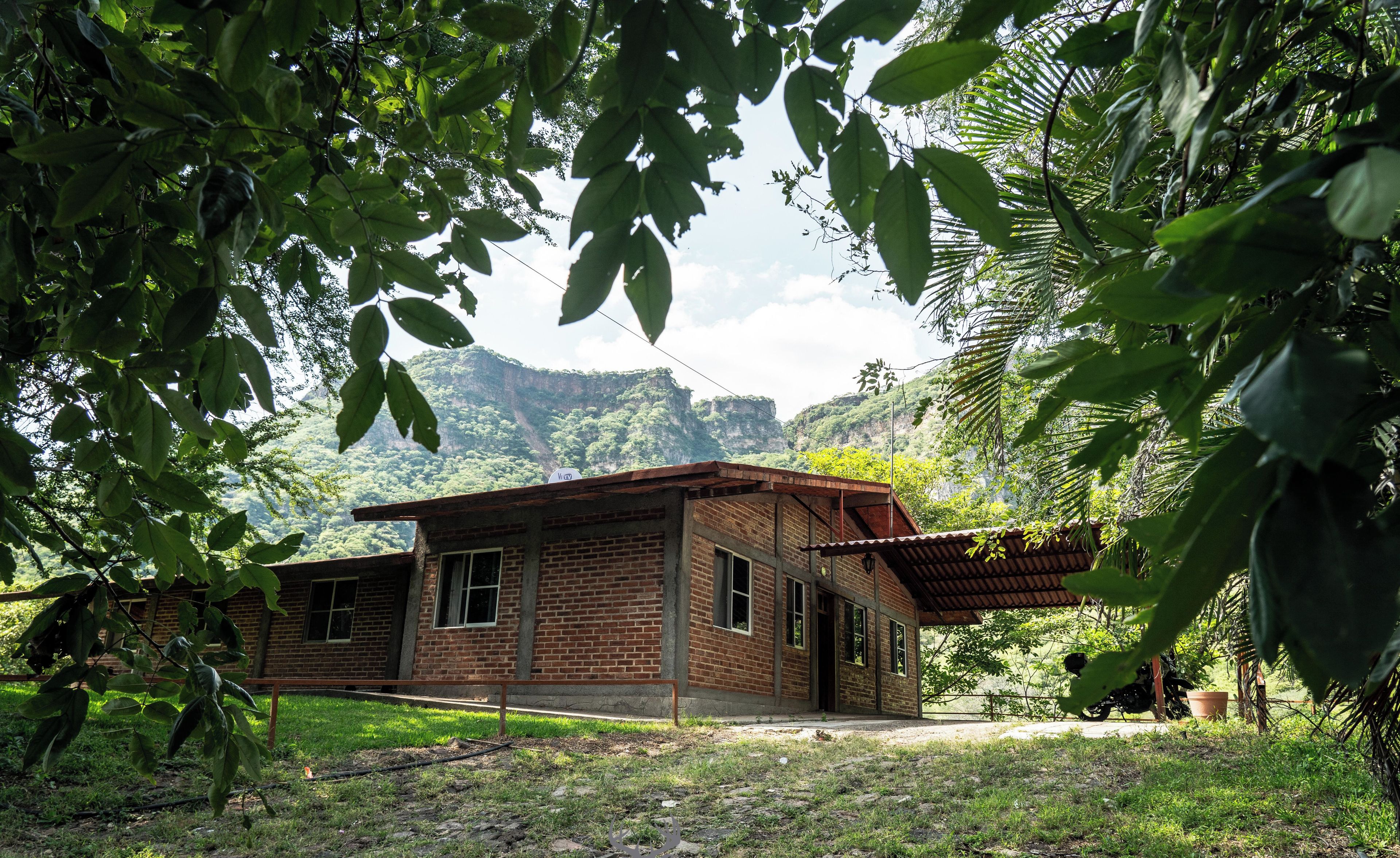 Edificio de ladrillo rústico de una sola planta, rodeado de exuberante vegetación y un paisaje montañoso.