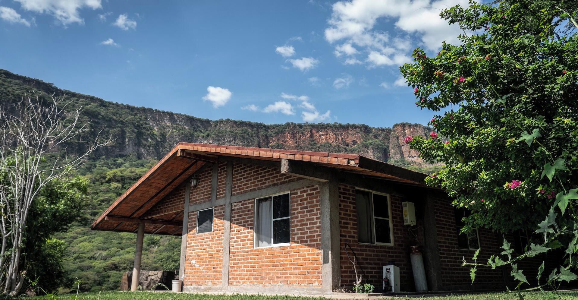 A small brick building with a sloped roof, a window, and a door, surrounded by greenery and a mountainous backdrop.