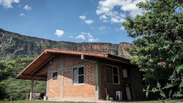 A small brick building with a sloped roof, a window, and a door, surrounded by greenery and a mountainous backdrop.