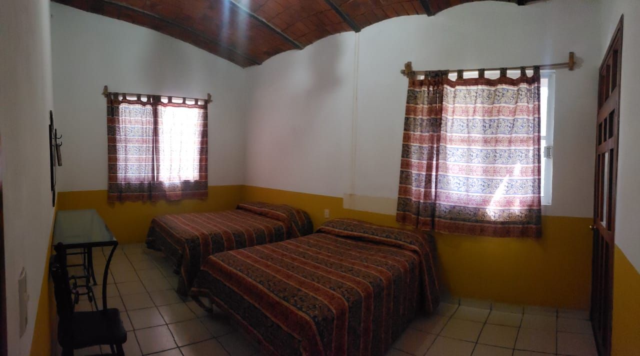 A room with two beds, a small table, and two windows with patterned curtains.