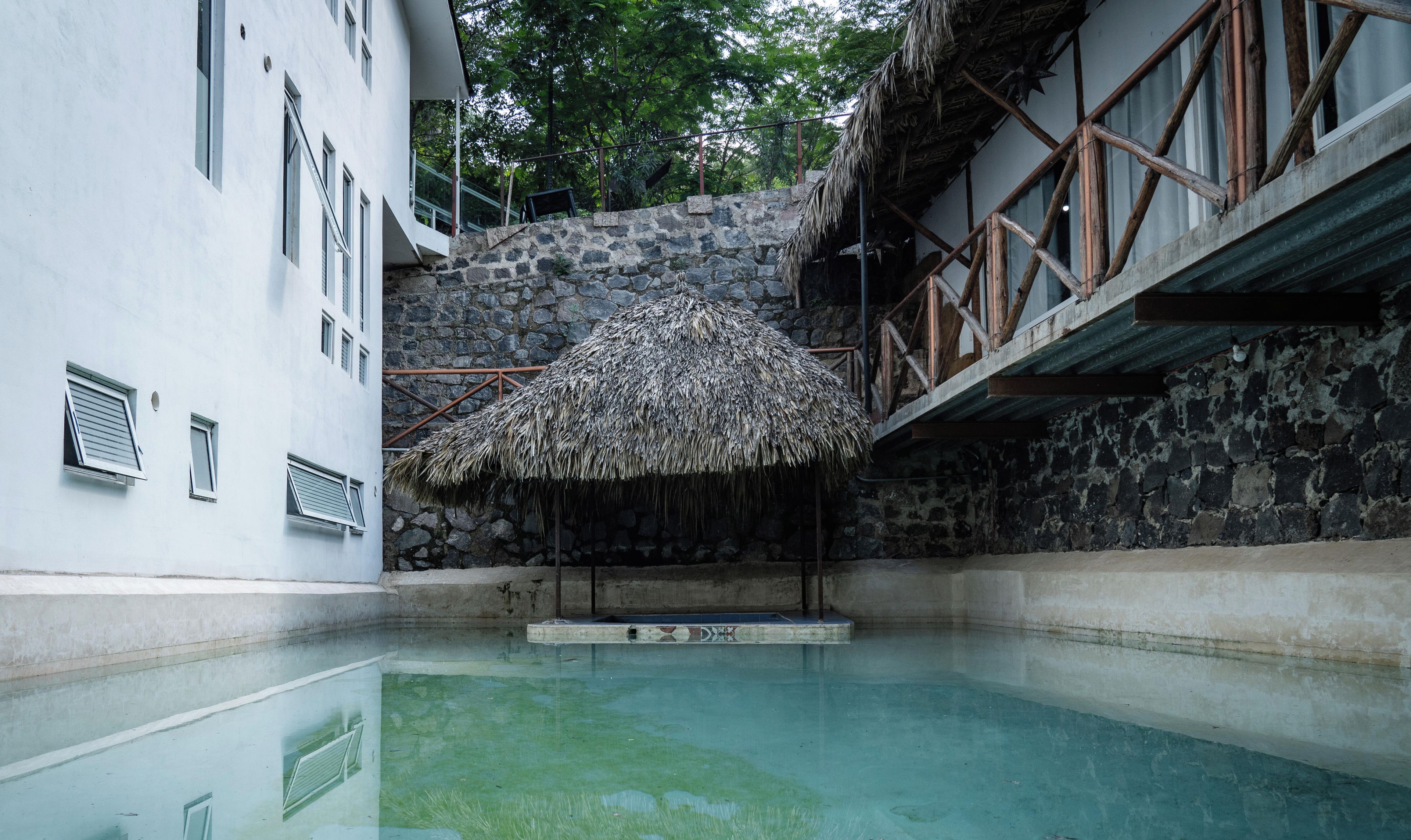 A swimming pool with a thatched roof structure, a stone wall, and a white building with windows.